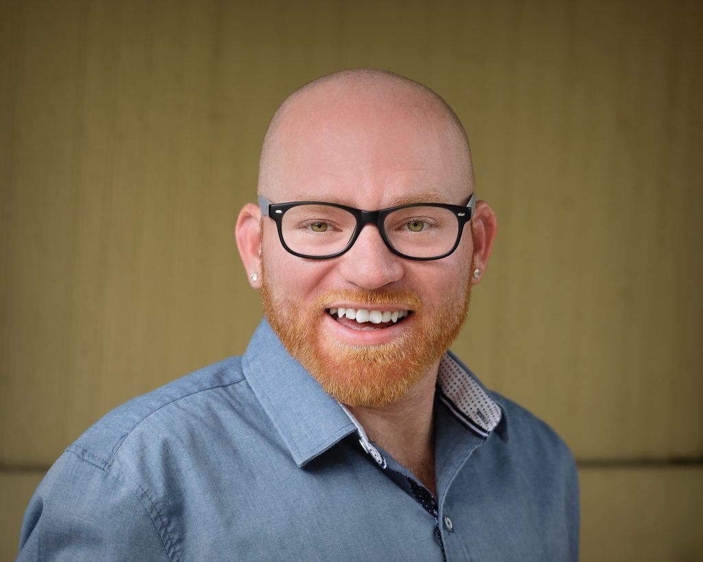 A photo of a bald man in a blue dress shirt, with glasses and a red beard smiling at the camera.